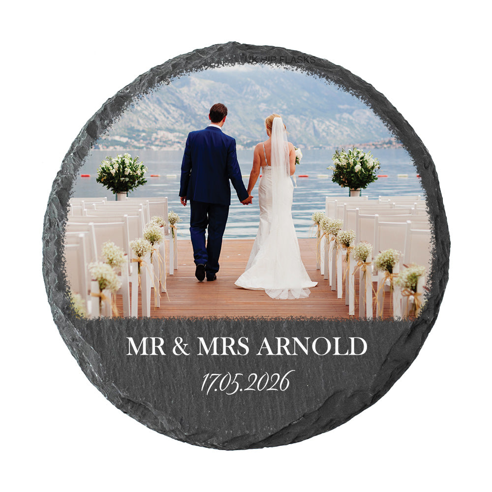 A round slate coaster featuring a custom wedding photo of a bride and groom holding hands on a dock with mountains and water in the background, personalized with the text 'MR & MRS ARNOLD' and the date '17.05.