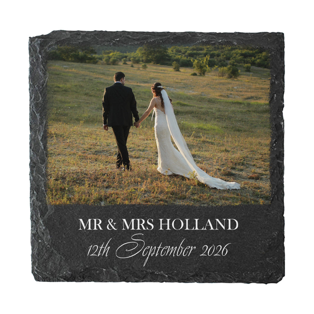 Square black slate wedding keepsake coaster featuring a custom photo of a bride and groom walking in a field, with personalized text reading 'MR & MRS HOLLAND 12th September 2026' on a white background.