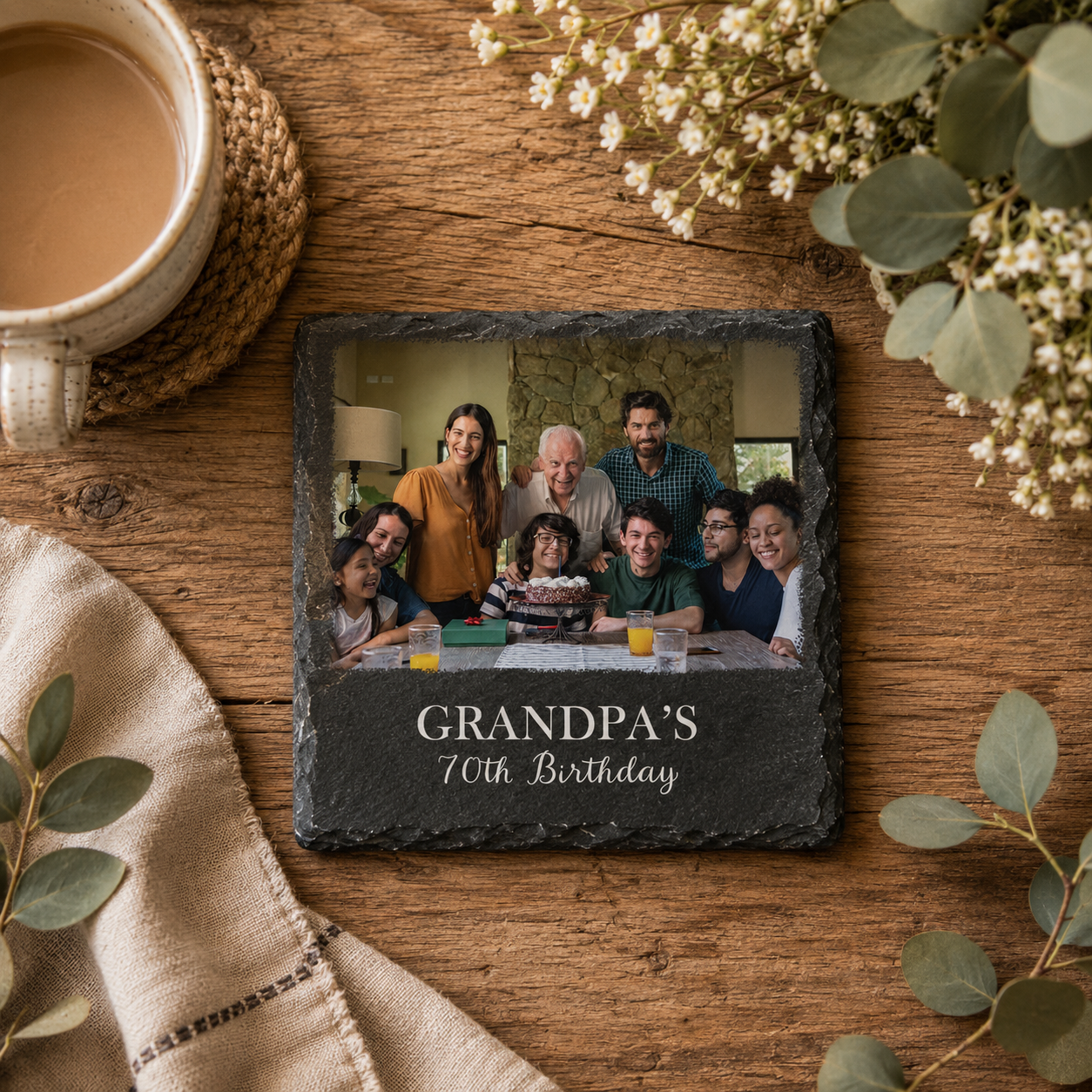 A square dark slate photo coaster on a rustic wooden table, featuring a custom family photo of a grandpa and his grandchildren with the text 'GRANDPA'S 70th Birthday'. The scene is styled with a cup of coffee, a woven coaster, a beige napkin, and eucalyptus leaves.