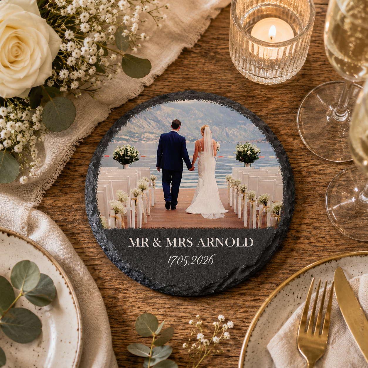 A round slate wedding coaster displayed on a rustic wooden table surrounded by plates, a candle, flowers, and wine glasses. The coaster features a custom printed photo of a bride and groom standing at the altar by the water, with the text 'MR & MRS ARNOLD' and the date.