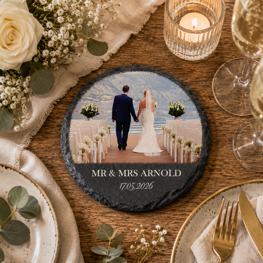 A round slate wedding coaster displayed on a rustic wooden table surrounded by plates, a candle, flowers, and wine glasses. The coaster features a custom printed photo of a bride and groom standing at the altar by the water, with the text 'MR & MRS ARNOLD' and the date.