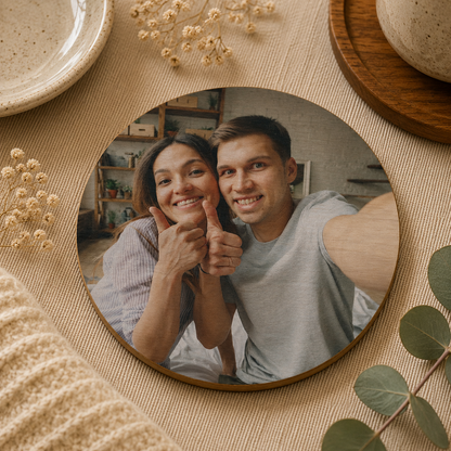A round wooden coaster personalised with a photo of a couple smiling, on a neutral surface surrounded by plants.