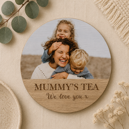 Round wooden coaster with a photo of a family and personalised text, surrounded by greenery and flowers on a beige background.
