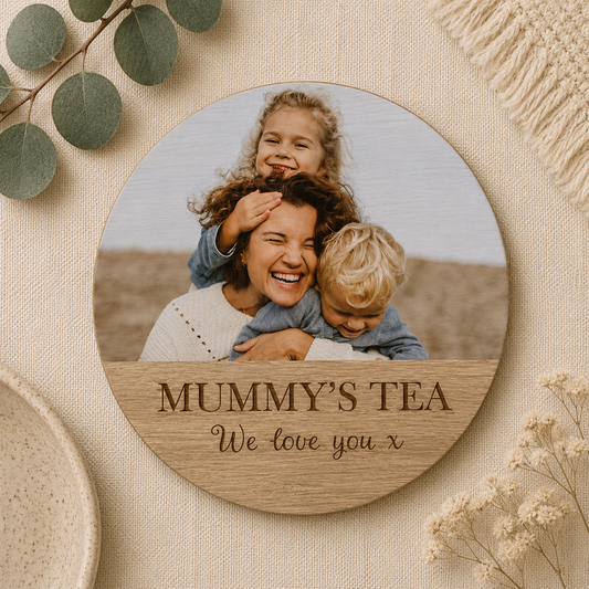 Round wooden coaster with a photo of a family and personalised text, surrounded by greenery and flowers on a beige background.
