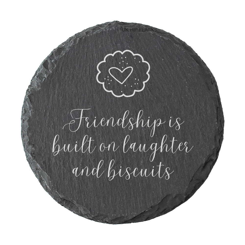 Round slate coaster with a heart and text reading "Friendship is built on laughter and biscuits" on a white background