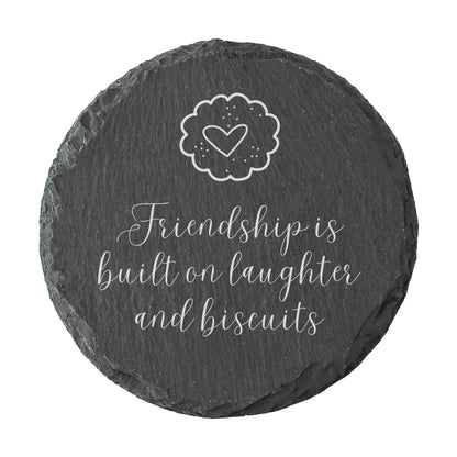 Round slate coaster with a heart and text reading "Friendship is built on laughter and biscuits" on a white background