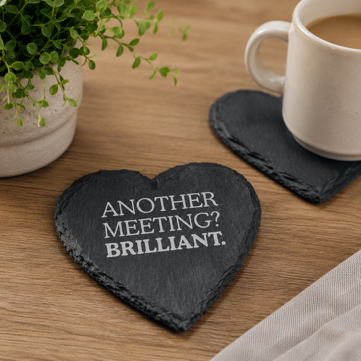 Heart-shaped slate coaster with 'Another meeting? Brilliant.' text on a wooden table with a mug and plant.