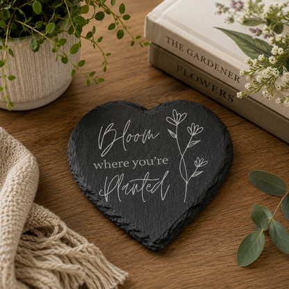 Heart-shaped slate with 'Bloom where you're planted' text on a wooden surface with plants and books.