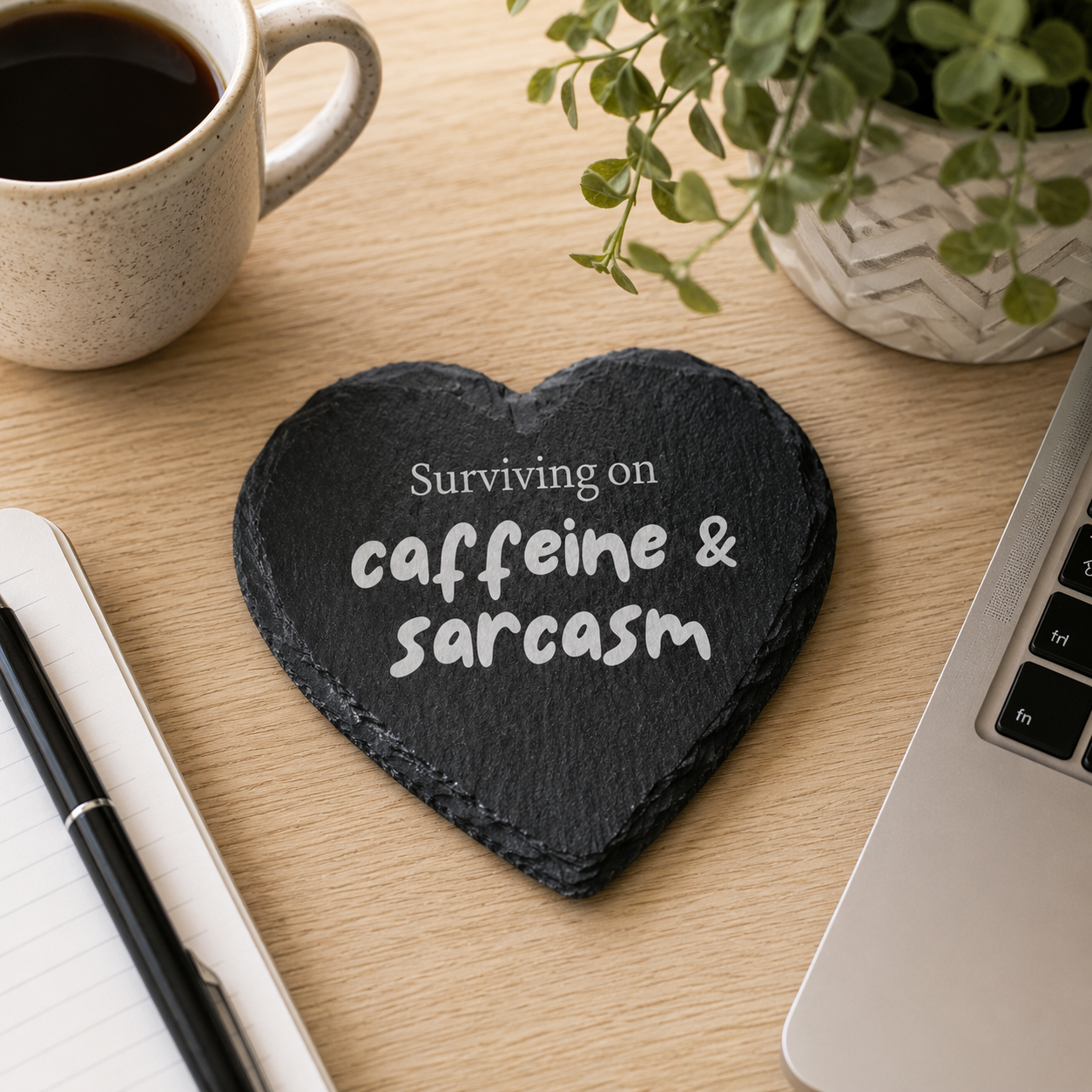 Heart-shaped slate coaster with 'Surviving on caffeine & sarcasm' text on a desk with coffee and laptop.