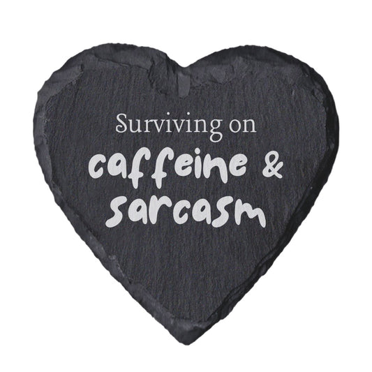 Heart-shaped slate coaster with text 'Surviving on caffeine & sarcasm' on a white background