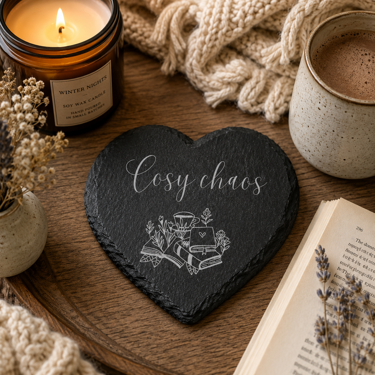 Heart-shaped slate coaster with 'Cosy chaos' text, lit candle, mug, and book on a wooden surface.