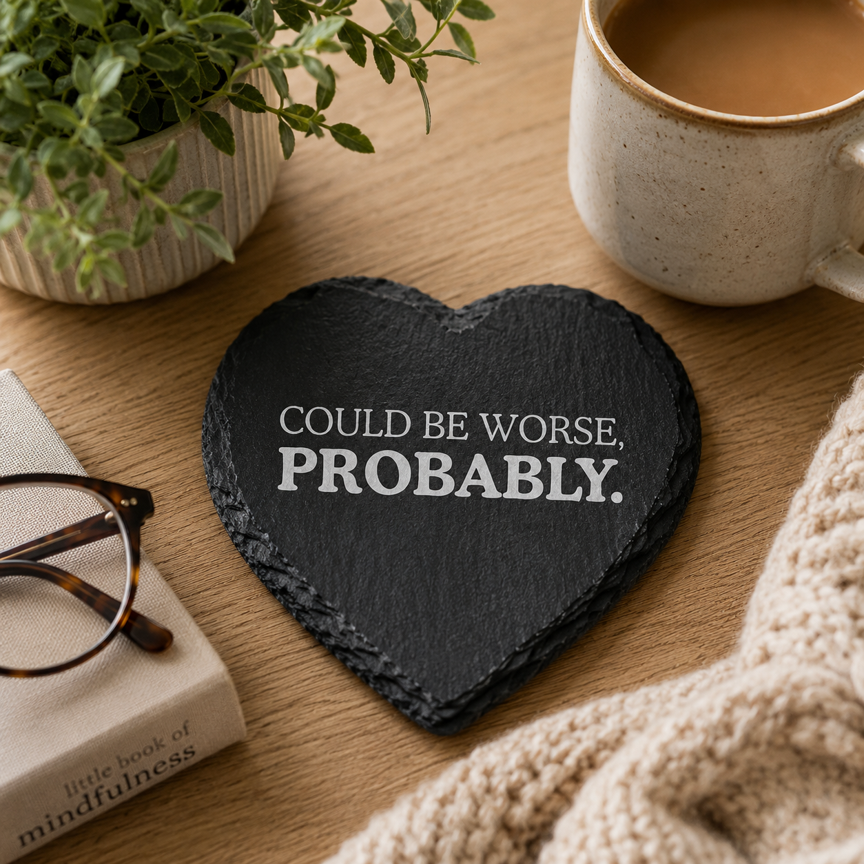 Heart-shaped slate coaster with 'Could be worse, probably' text on a wooden table with a mug, glasses, and book.