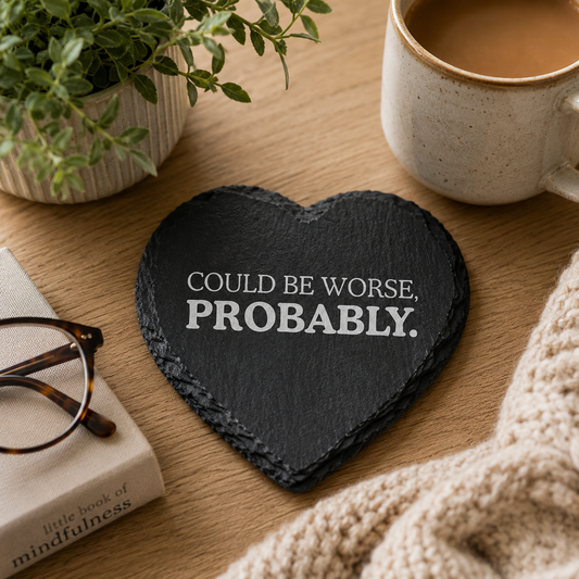Heart-shaped slate coaster with 'Could be worse, probably' text on a wooden table with a mug, glasses, and book.