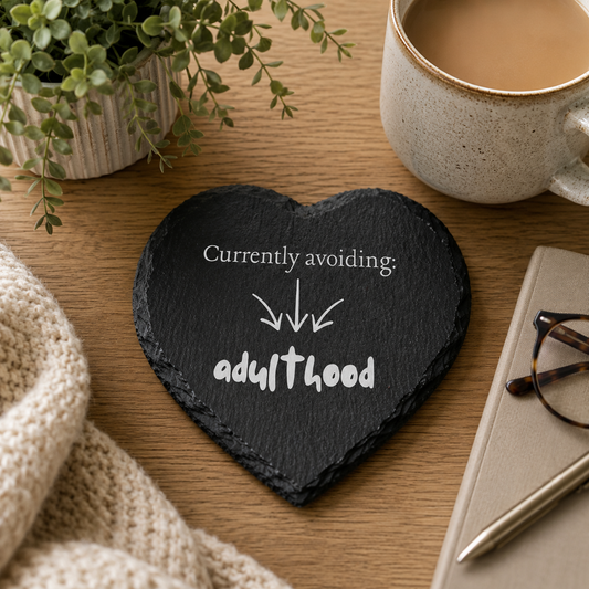 Heart-shaped slate coaster with text 'Currently avoiding adulthood' on a wooden table with a cup of coffee, plant, and glasses.