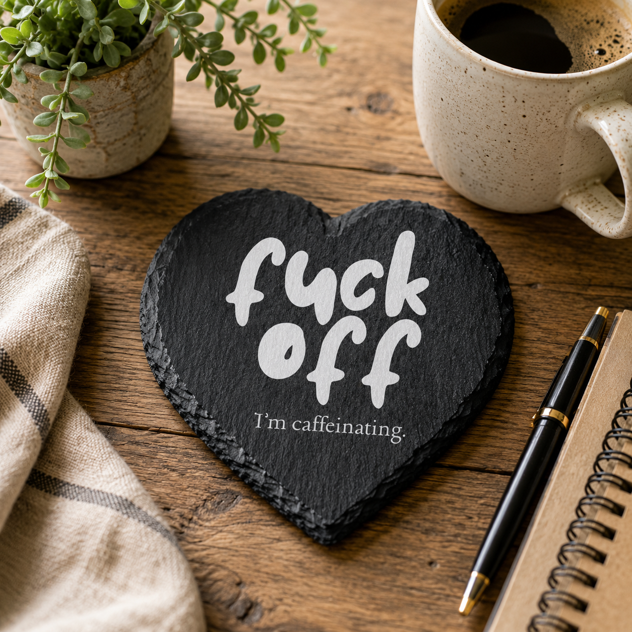 Heart-shaped slate coaster with 'fuck off, I'm caffeinating' text on a wooden surface with a mug and plant.