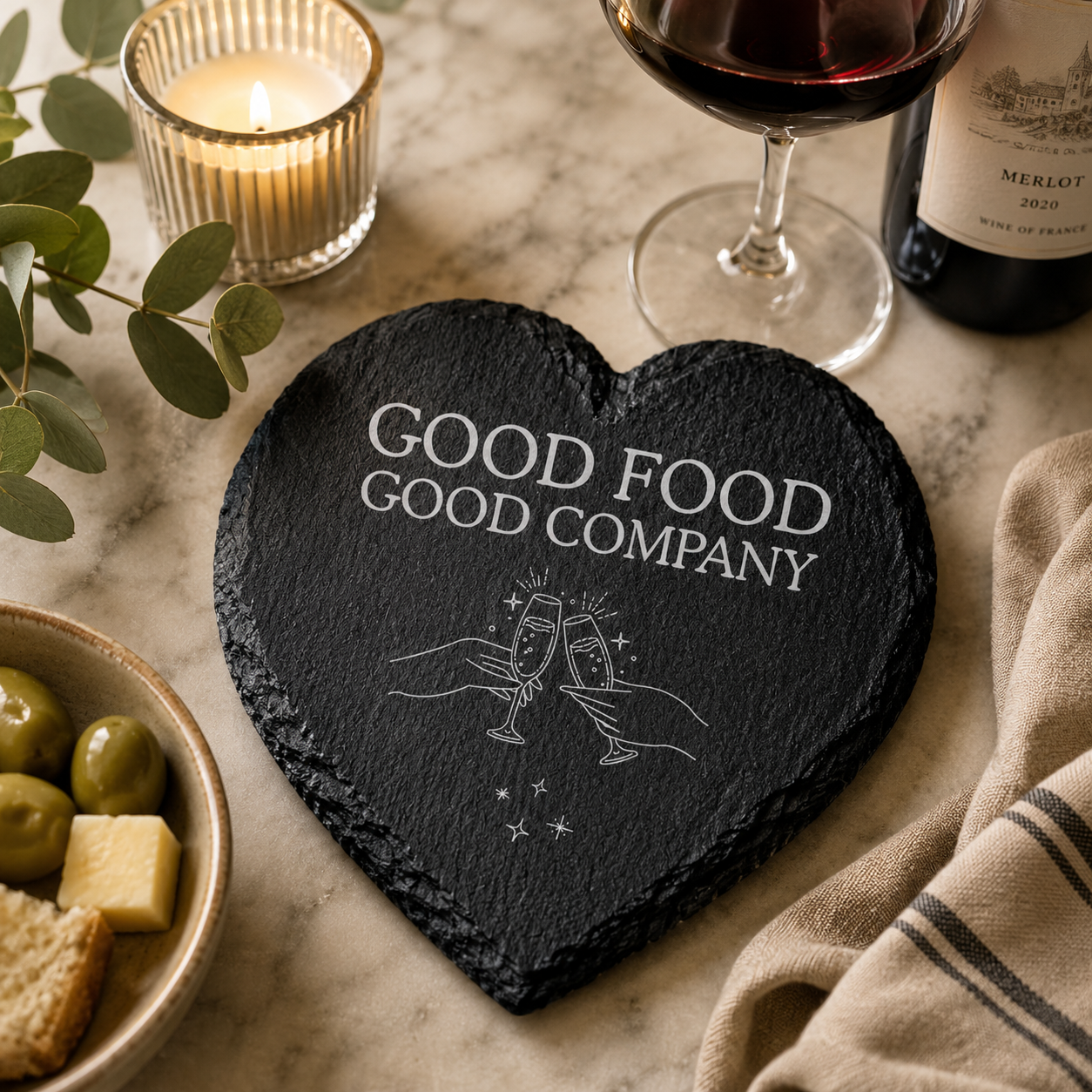 Heart-shaped slate coaster with 'Good Food Good Company' text on a table with wine and snacks.