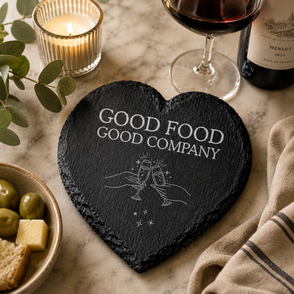 Heart-shaped slate coaster with 'Good Food Good Company' text on a table with wine and snacks.