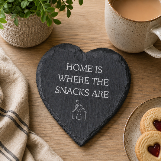 Heart-shaped slate coaster with 'Home is where the snacks are' text on a wooden table with a mug, cookies, and plant.