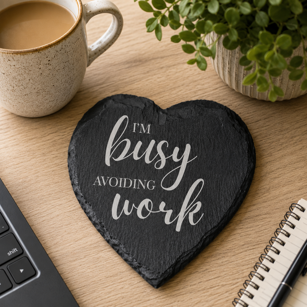Heart-shaped slate coaster with 'I'm busy avoiding work' text on a desk with a laptop, coffee, and plant.