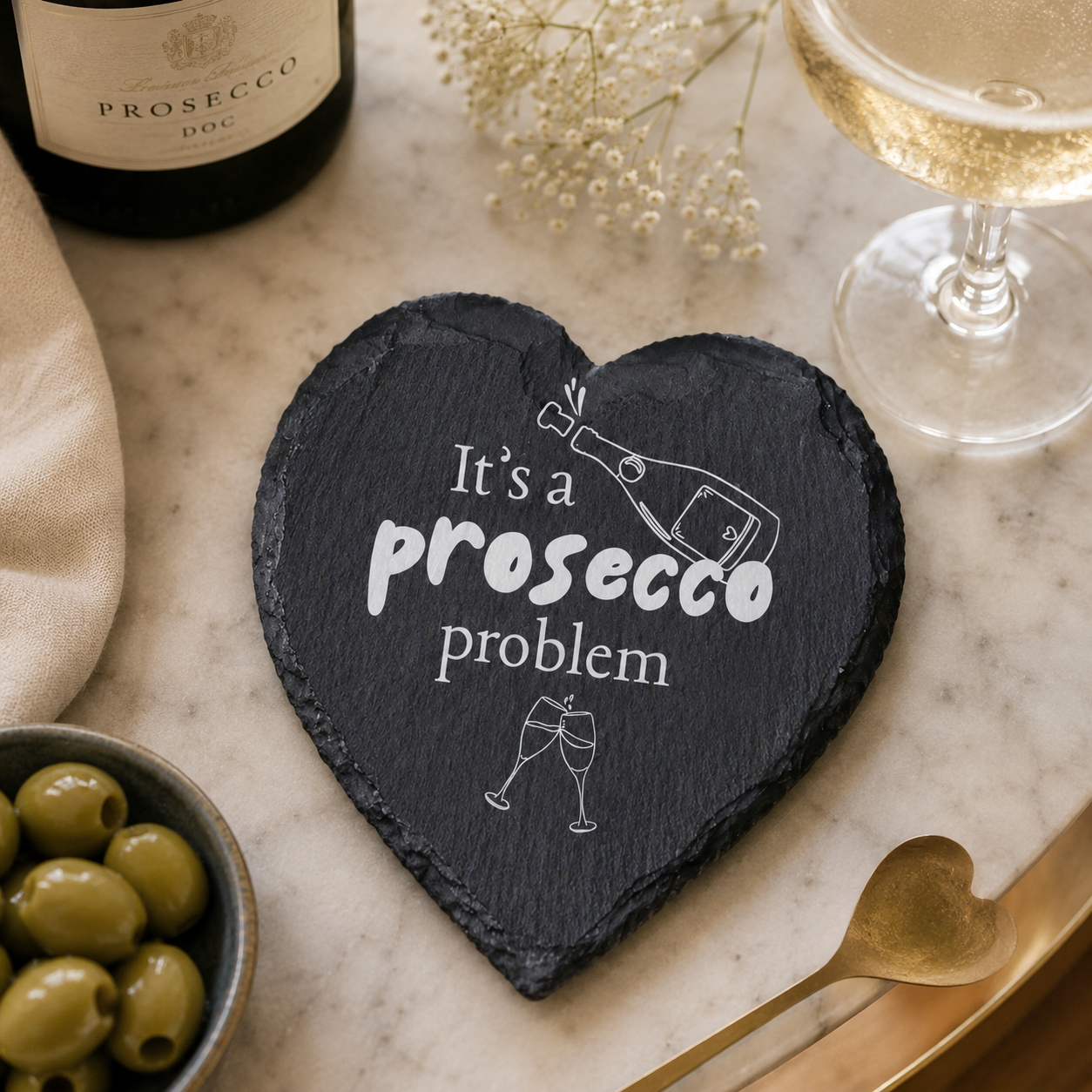 Heart-shaped slate coaster with 'It's a prosecco problem' text on a marble surface with wine and olives.