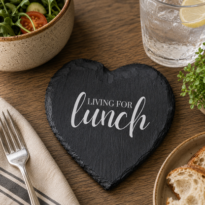 Heart-shaped slate coaster with 'Living for lunch' text on a dining table with salad, bread, and water.