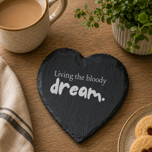 Heart-shaped slate coaster with 'Living the bloody dream' text on a wooden table with a cup of coffee and cookies.