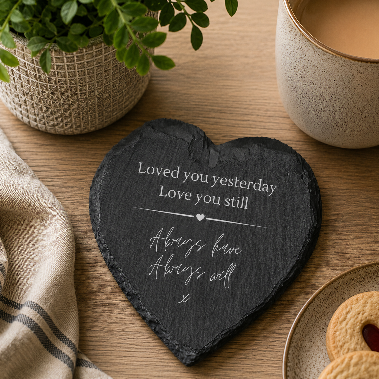 Heart-shaped slate coaster with white text on a wooden table with a cup of coffee and cookies.