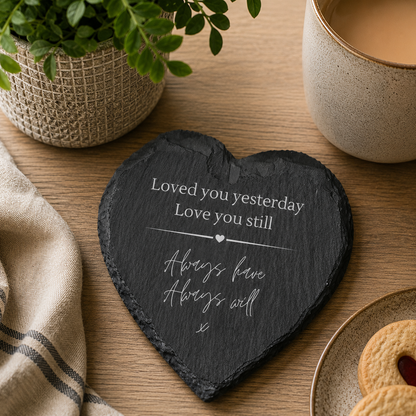 Heart-shaped slate coaster with white text on a wooden table with a cup of coffee and cookies.