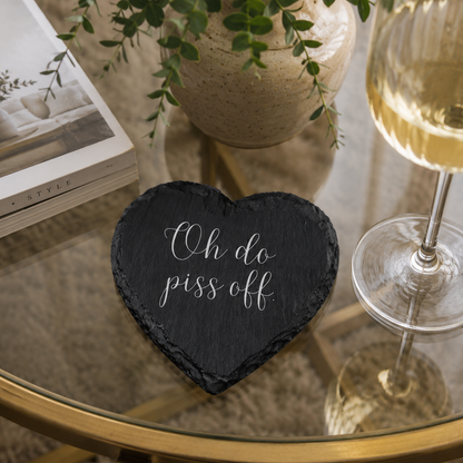 Heart-shaped slate coaster with "oh do piss off" in white text on a glass table with a wine glass and book.