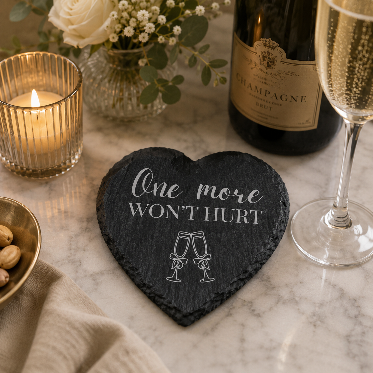 Heart-shaped slate coaster with 'One more WON'T HURT' text on a marble surface with champagne, candles, and flowers.