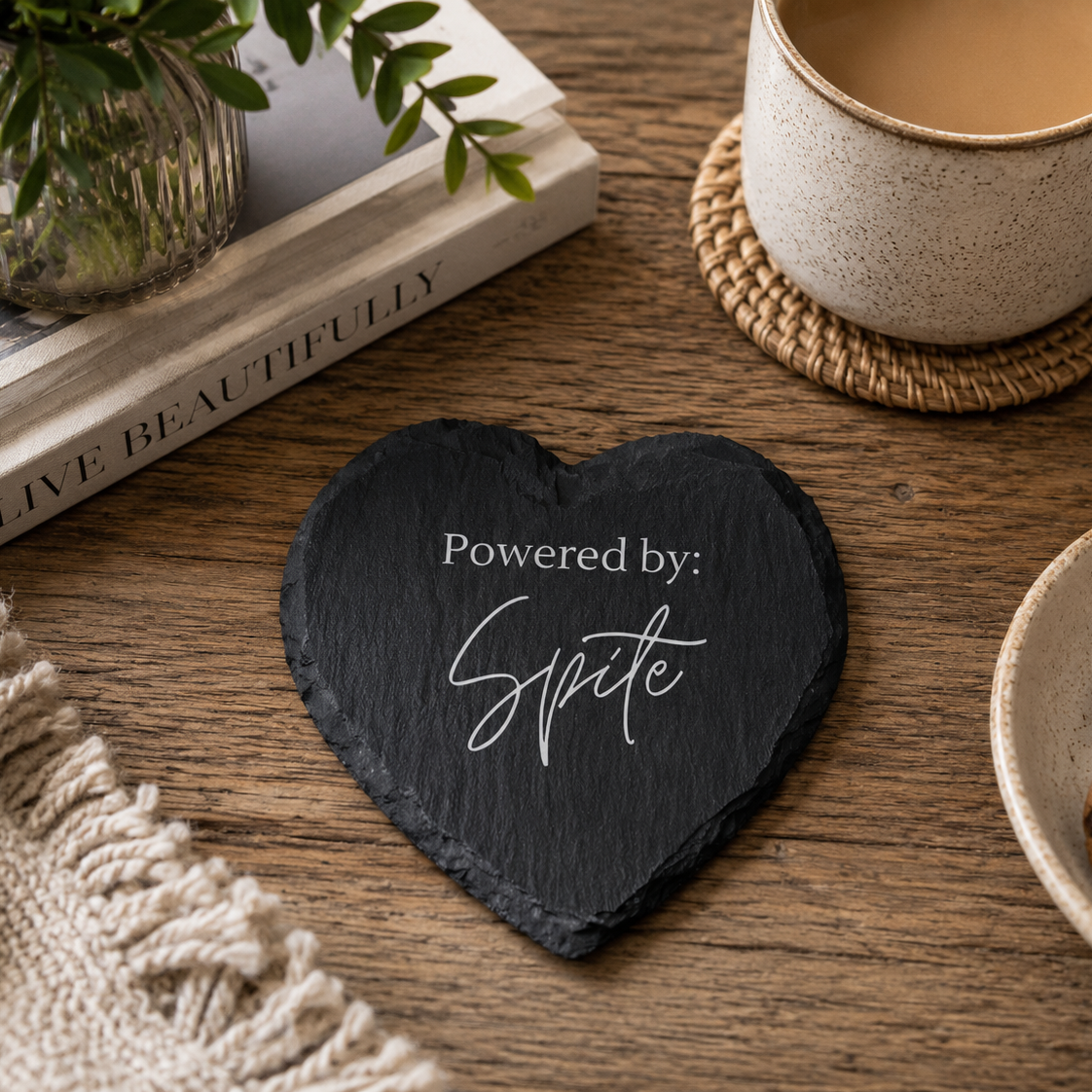 Heart-shaped slate coaster with 'Powered by: Spite' text on a wooden table with a cup of coffee, book, and cookies.