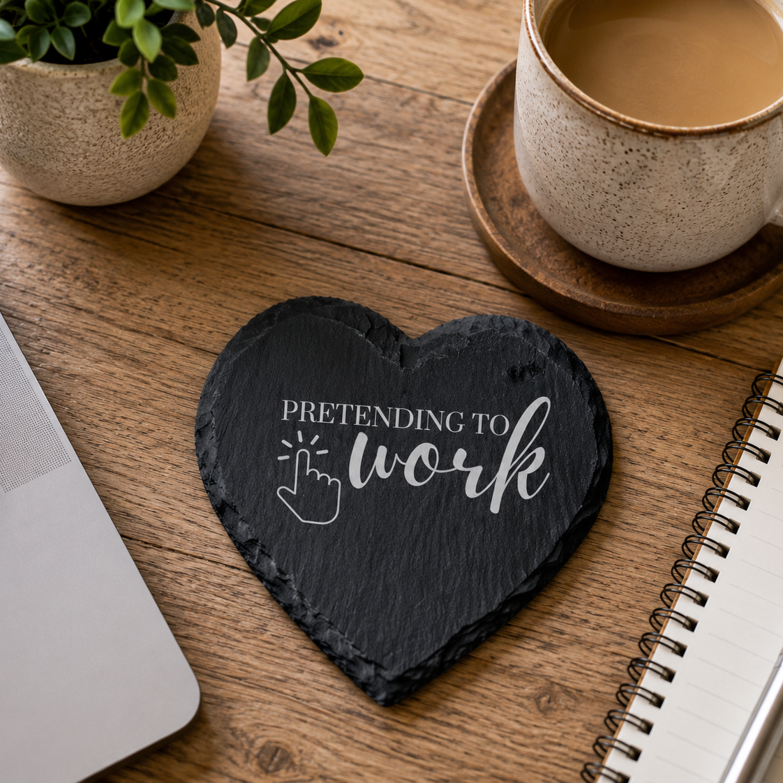 Heart-shaped slate coaster with 'Pretending to work' text on a wooden desk with laptop, coffee, and notebook.