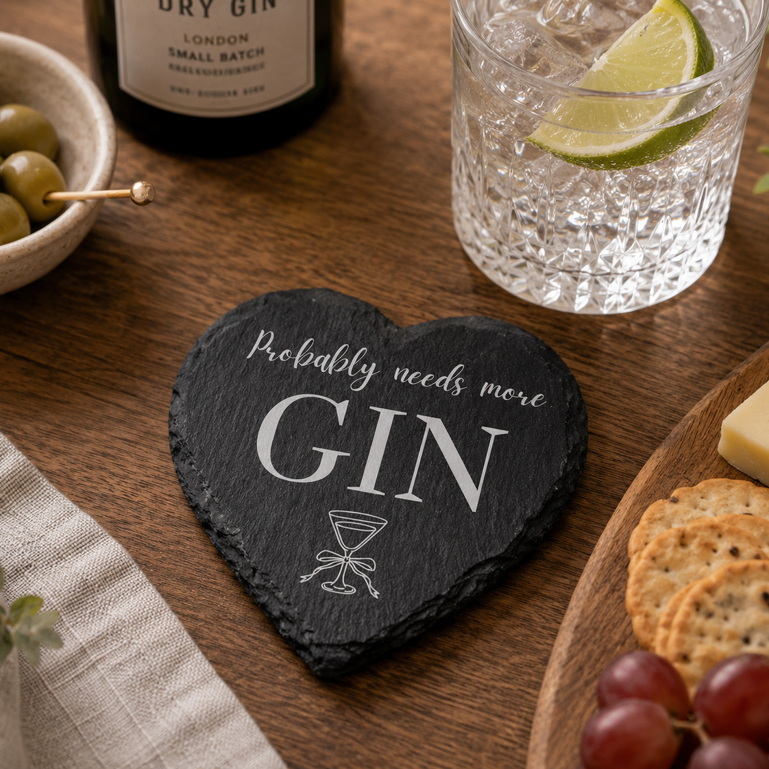 Heart-shaped slate coaster with 'Probably needs more Gin' text on a table with gin and tonic, olives, and cheese.