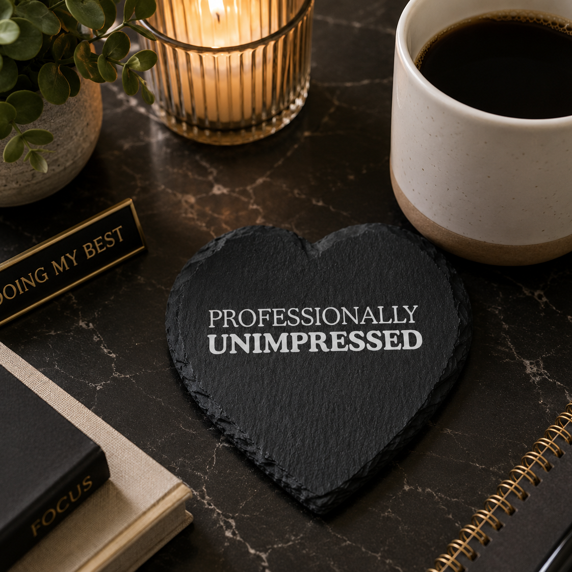 Heart-shaped slate coaster with 'PROFESSIONALLY UNIMPRESSED' text on a marble desk with coffee and office items.