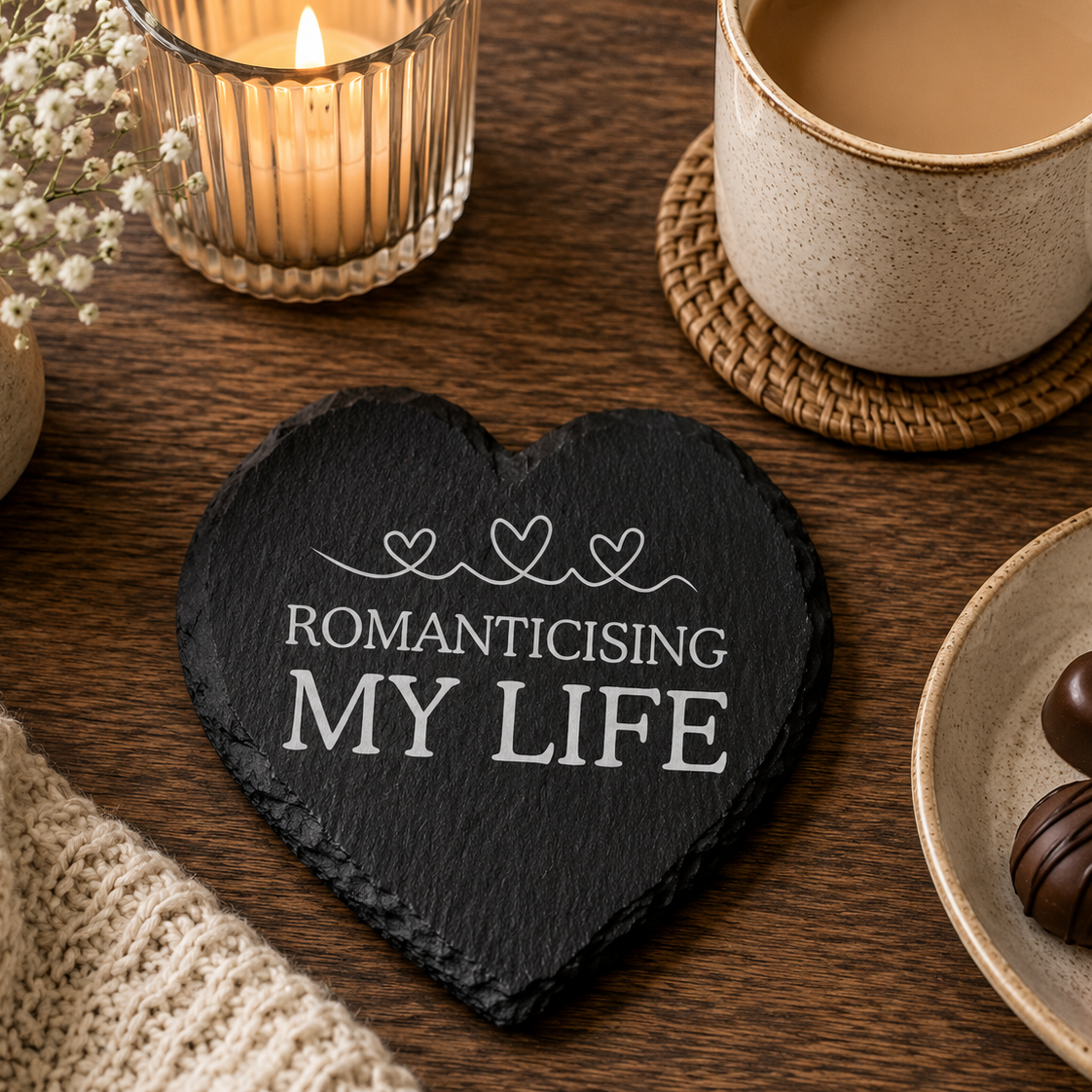 Heart-shaped slate coaster with 'Romanticising My Life' text on a table with coffee, chocolates, and flowers.