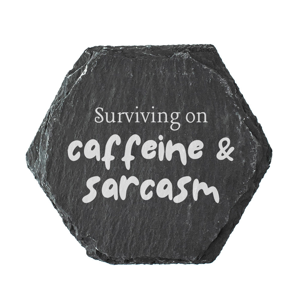 Hexagonal black slate coaster with 'Surviving on caffeine & sarcasm' text on a white background
