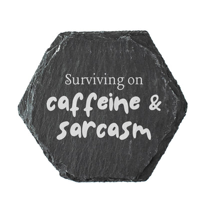 Hexagonal black slate coaster with 'Surviving on caffeine & sarcasm' text on a white background