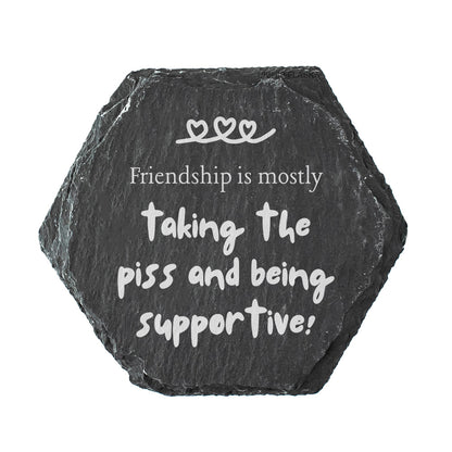 Hexagonal black slate coaster with white text 'Friendship is mostly taking the piss and being supportive!' on a white background