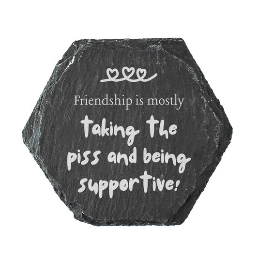 Hexagonal black slate coaster with white text 'Friendship is mostly taking the piss and being supportive!' on a white background