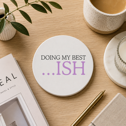 A round white ceramic coaster with the quote 'Doing my best... ISH' in grey and purple text. The coaster is set on a wooden desk with tea and stationery.