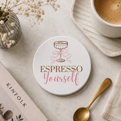 Round ceramic coaster featuring the witty quote "Espresso yourself" in warm brown and pink text, topped with an illustrated espresso martini glass. The coaster is on a stone surface with a coffee cup and teaspoon.