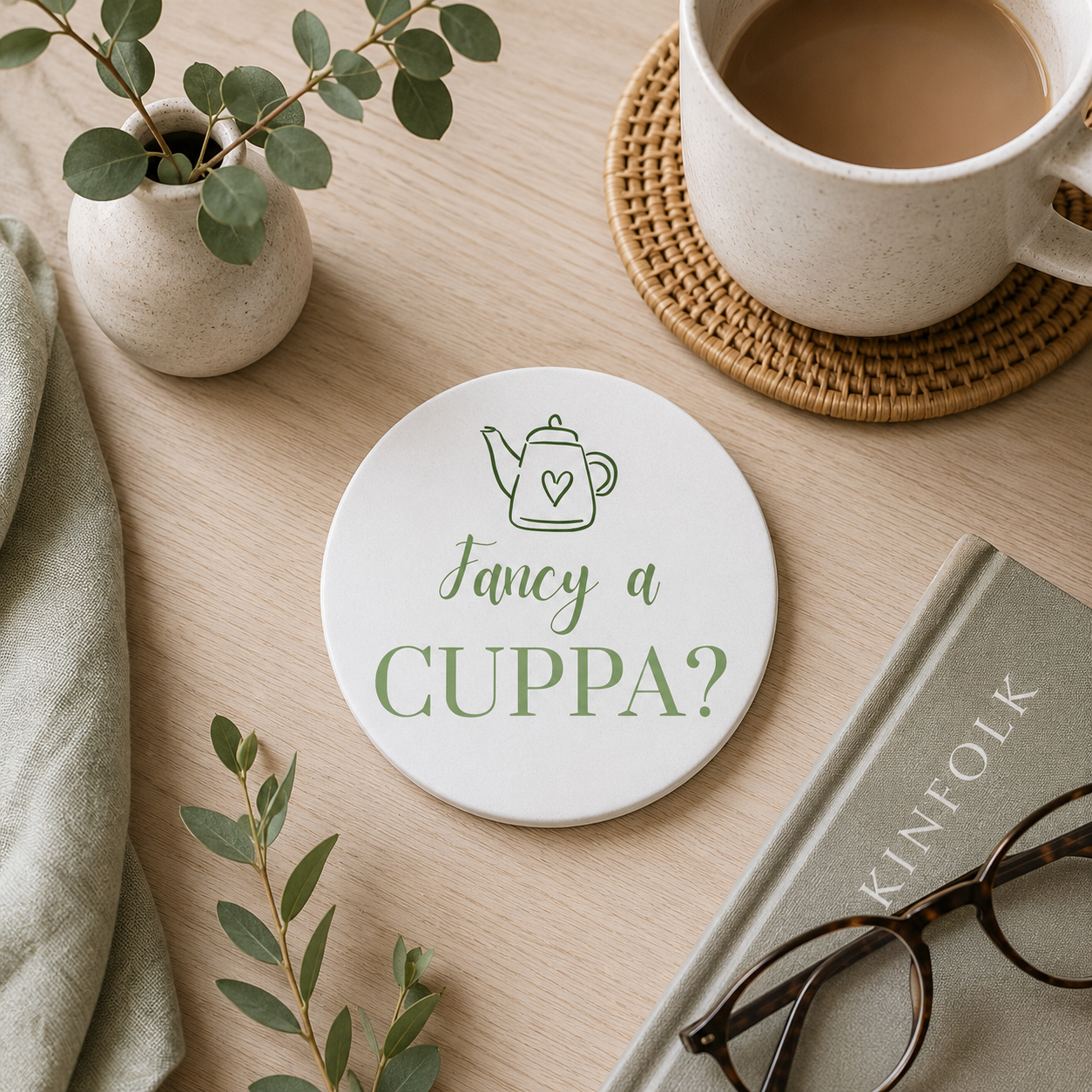 Round ceramic coaster featuring the cheerful quote "Fancy a cuppa?" in crisp green lettering. The coaster is on a wooden table with a cup of tea and books.