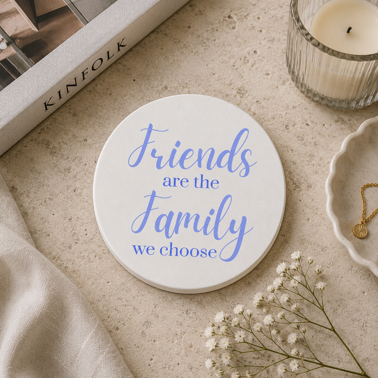 Round ceramic coaster featuring the thoughtful quote "Friends are the family we choose" in crisp lilac lettering. The coaster is on a stone surface with candles and books.