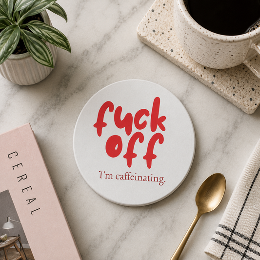 Round ceramic coaster featuring the humorous quote "Fuck off I'm caffeinating" in crisp red lettering. The coaster is on a marble surface with a coffee and spoon.