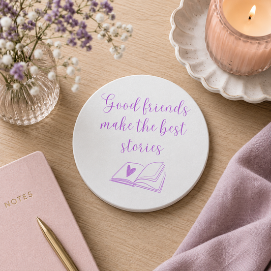 Round ceramic coaster on a wooden surface with candle and notebook, the coaster features the friendship quote "Good friends make the best stories" in crisp purple lettering with a book illustration underneath.
