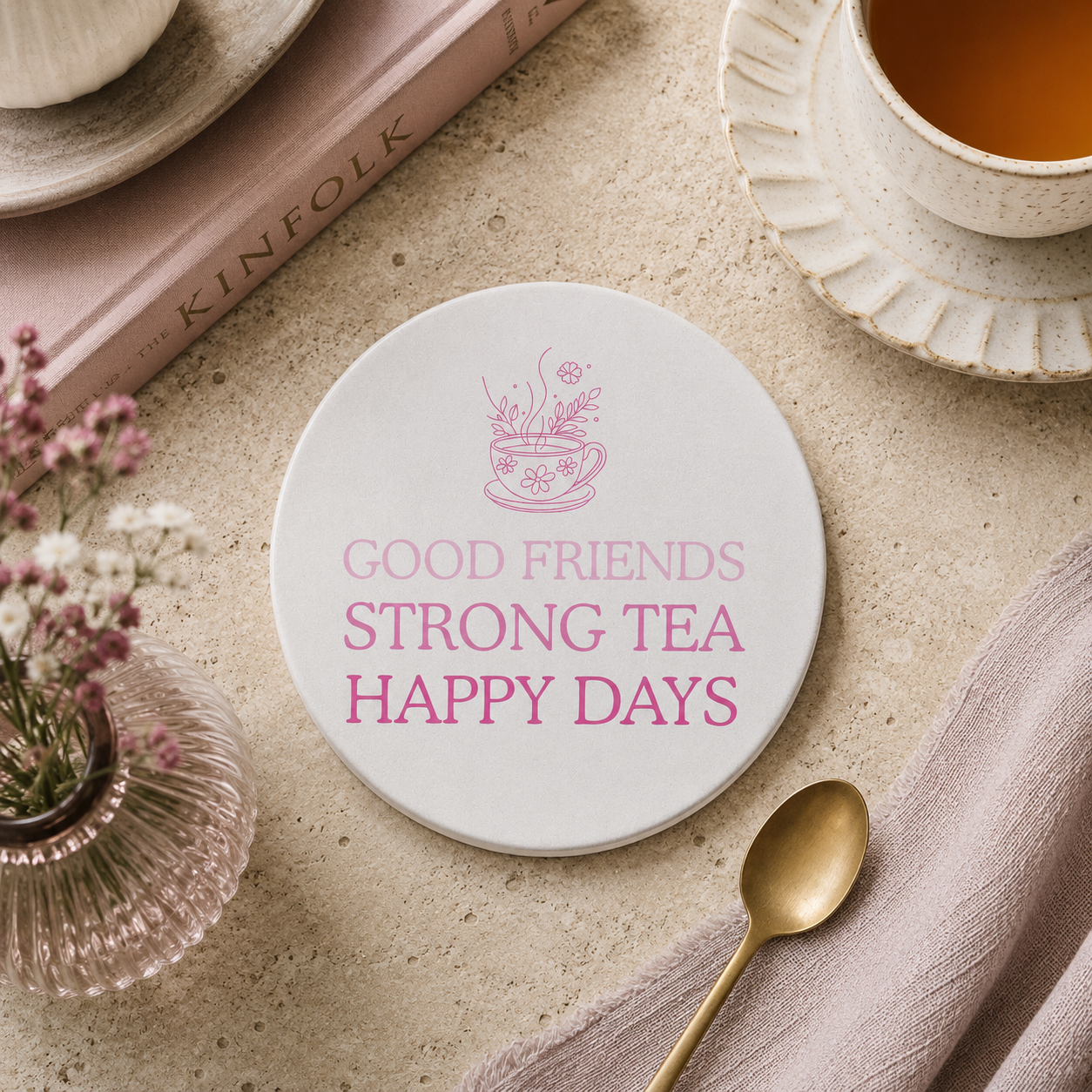 Round ceramic coaster featuring the fun quote "Good friends, strong tea, happy days" in crisp pink lettering with a teacup illustration. The coaster is on a table with a cup of tea and plants.