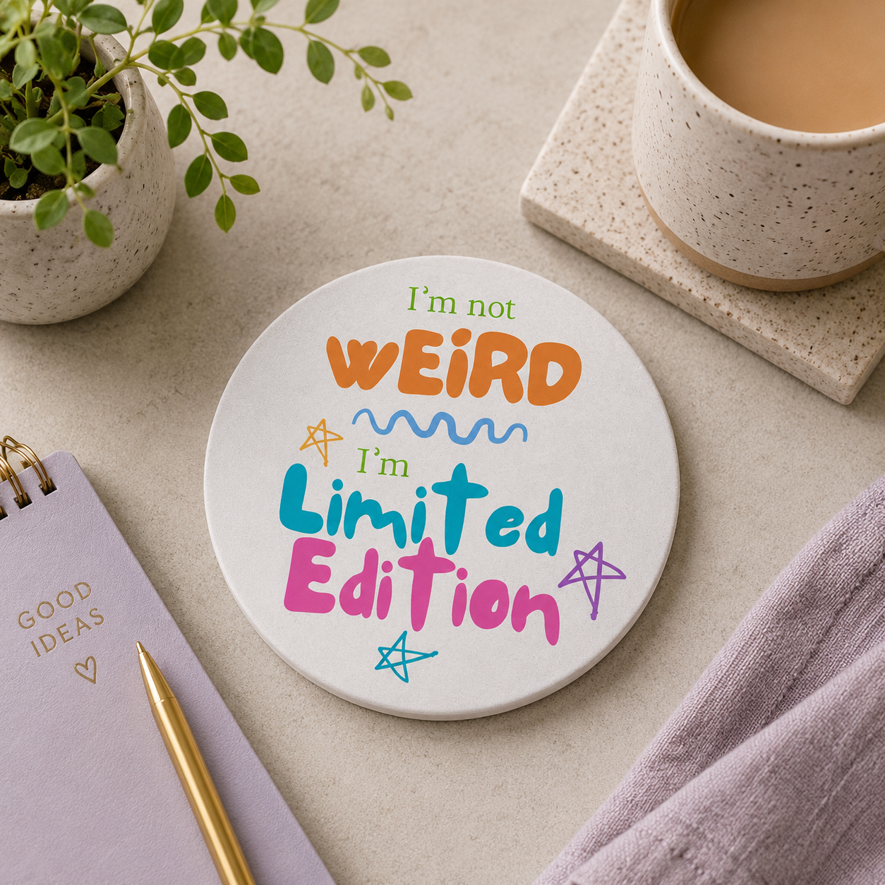 Round ceramic coaster featuring the quirky quote "I'm not weird, I'm limited edition" in colourful lettering. The coaster is on a desk with a cup of tea and notebook.