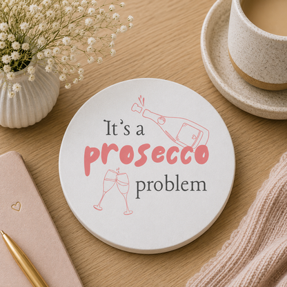 Round ceramic coaster featuring the humorous quote "It's a prosecco problem" in crisp pink lettering. The coaster is on a wooden table with a cup of tea and notebook. 