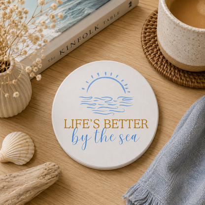 Round ceramic coaster featuring the quote "Life's better by the sea" in blue and gold lettering and a sun and sea illustration. The coaster is on a wooden table in a coastal theme room.