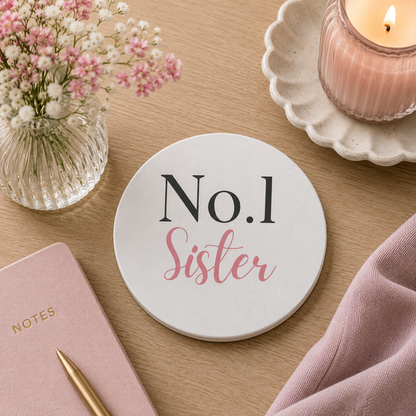 Round ceramic coaster features "No. 1 Sister" in dark grey and pink text. The coaster is on a wooden surface with a candle and notebook.
